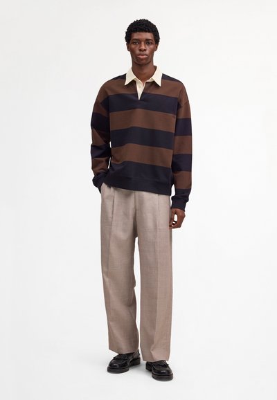 Homme debout portant un maillot de rugby à rayures marron et noir, un pantalon large beige et des mocassins en cuir noir sur un fond blanc uni.