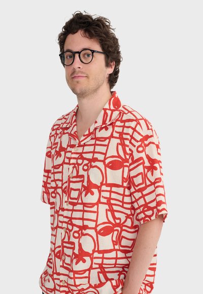 Homme aux cheveux bouclés et lunettes portant une chemise à manches courtes à boutons avec un motif abstrait de fruits rouges et une grille sur fond blanc.