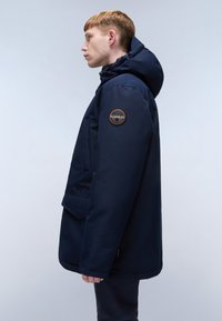 Giacca cappuccio blu navy con texture liscia, dotata di tasca frontale e una toppa rotonda con logo sulla manica sinistra. Vista laterale mostrata.