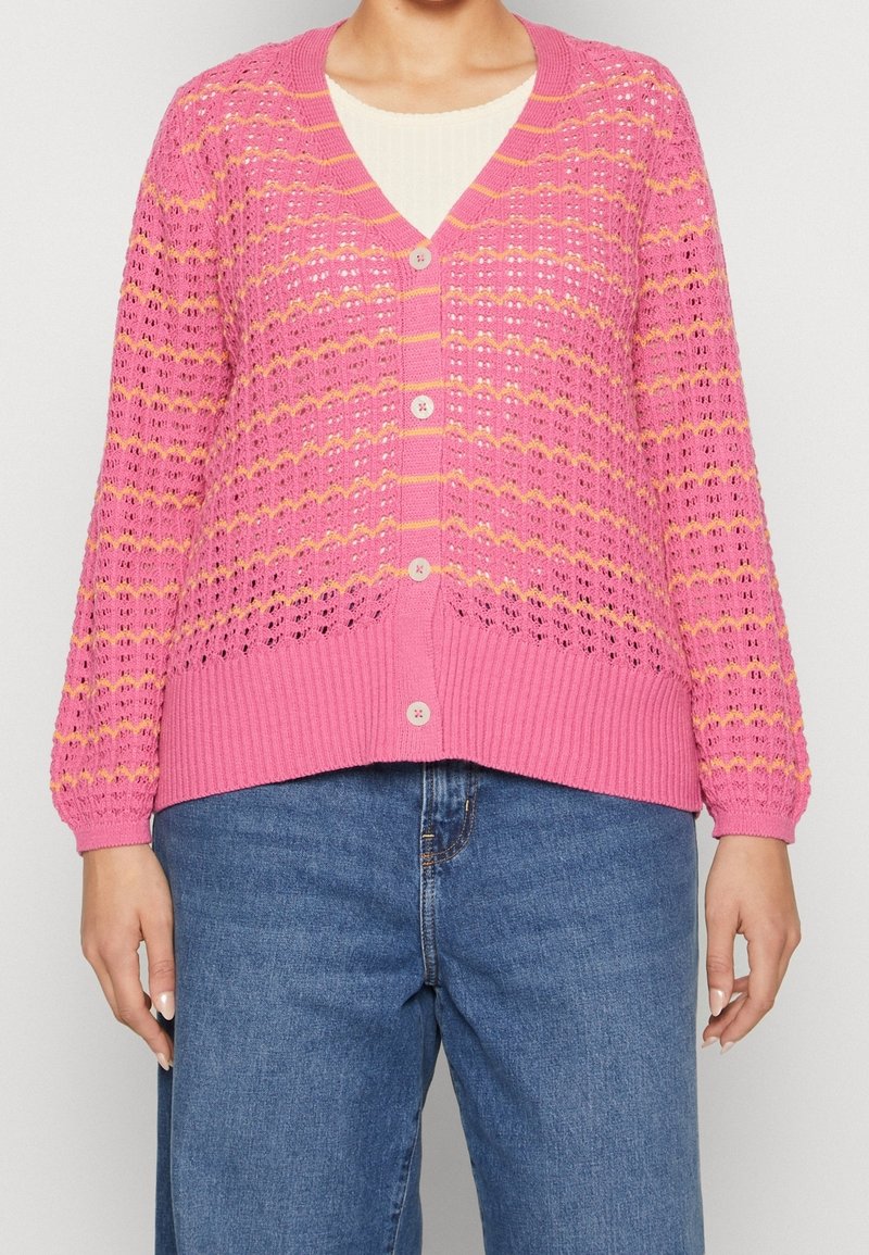Femme portant un cardigan en tricot rose à boutons avec des rayures jaunes par-dessus un haut blanc et un jean bleu.