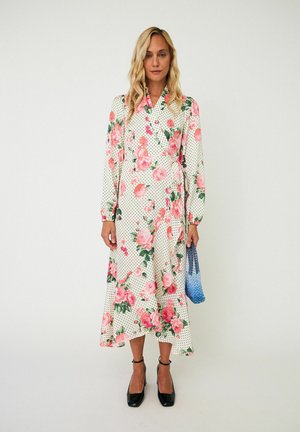 Femme portant une longue robe blanche avec un motif floral rose et pois noirs, des talons noirs, tenant un petit sac à main bleu texturé.