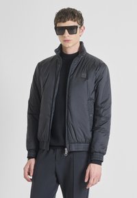 Giacca bomber nera in nylon con zip frontale, polsini e bordo ribatti. Presenta tasche laterali e un discreto logo del marchio sul petto.