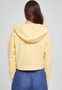 Felpa gialla a sacchetto con cappuccio regolabile, morbida al tatto e con polsini a coste, abbinata a una gonna blu.
