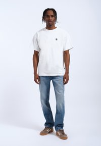 T-shirt bianco in cotone con un piccolo logo nero, abbinato a jeans in denim blu chiaro e sneakers in suede marroni. Design semplice e dalla vestibilità rilassata.