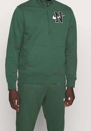 Mężczyzna ubrany w zielony dres Nike z pulowerem na półzamek, na którym znajduje się duża litera "N" oraz biały znak Nike na piersi i spodniach.