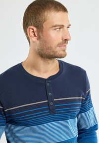 Homme avec des cheveux bruns clairs courts et une barbe, portant une chemise à manches longues bleu marine avec des rayures horizontales bleues et blanches, regardant vers la droite.