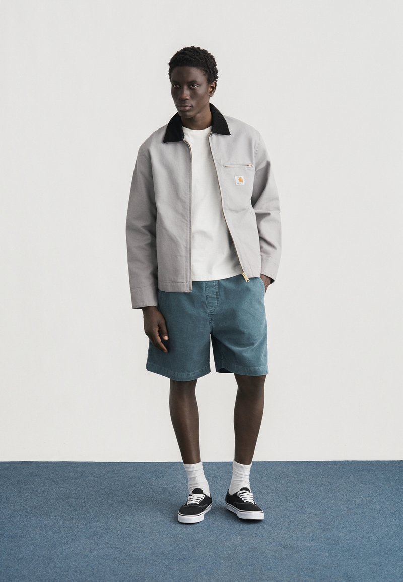 Jeune homme debout à l'intérieur, portant une veste grise, un t-shirt blanc, un short bleu, des chaussettes blanches et des baskets noires sur un tapis bleu.