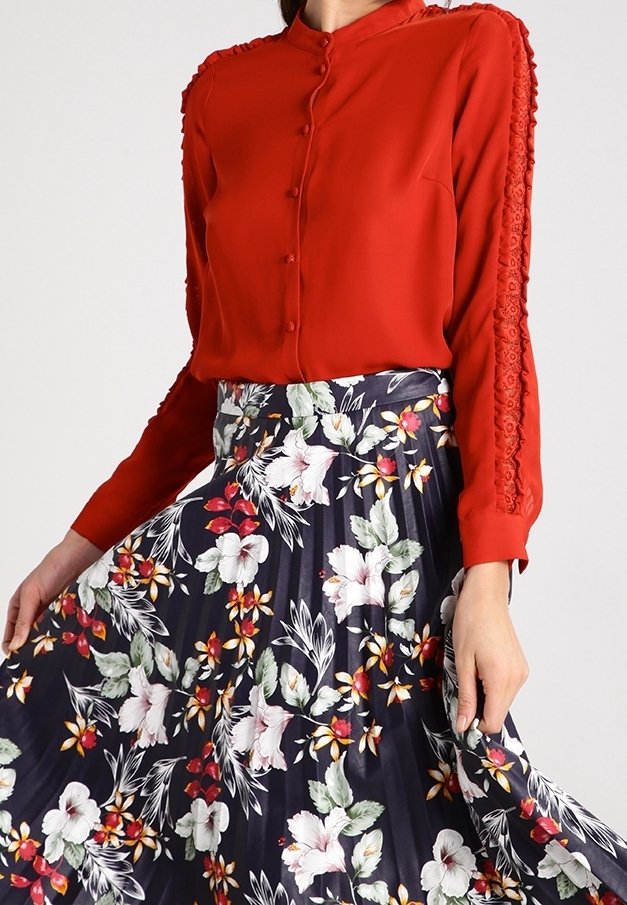 Femme portant un chemisier rouge à manches longues avec des boutons décoratifs et une jupe plissée bleu marine avec des motifs floraux blancs et rouges.