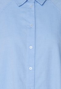 Chemise col bleu clair en tissu texturé, avec une rangée verticale de quatre boutons blancs sur le devant.