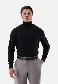 Ein Mann trägt einen schwarzen Rollkragenpullover, der in eine hellgraue Hose mit einem schwarzen Gürtel gesteckt ist, und steht vor einem schlichten Hintergrund.