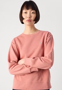 Young woman with short dark hair wears a pink crewneck sweatshirt, standing with arms crossed against a plain white background.