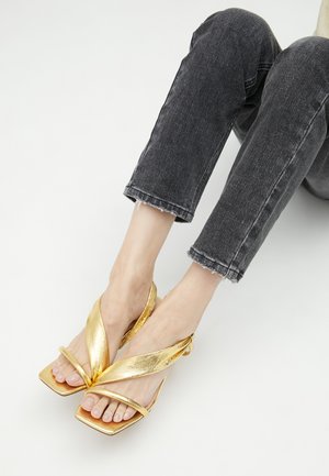 Sandalias metálicas doradas con un diseño de tiras cruzadas, punteras cuadradas y una suela plana. Combinadas con jeans negros cortos y deshilachados.