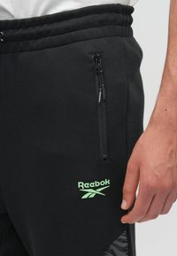 Čierne teplákové nohavice vyrobené z mäkkej tkaniny, s vreckom na zips a zeleným logom Reebok. Textúrovaná časť po stranách pridáva detail.