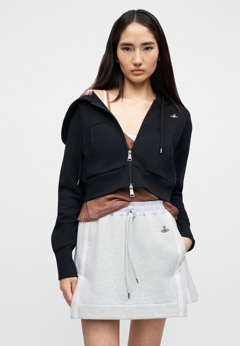 Sweat à capuche court noir zippé avec une doublure à carreaux dans la capuche, associé à une jupe grise à cordon de serrage avec des poches latérales et des rayures claires.