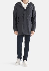 Marineblauer wasserdichter Parka mit Kapuze, ausgestattet mit Druckknöpfen, zwei Seitentaschen, über einem weißen Hoodie getragen, kombiniert mit dunklen Jeans und weißen Sneakers.