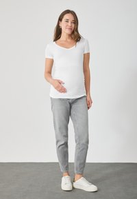 Balta, īsām piedurknēm, grūtniecību topa blūze ar V veida kakla izgriezumu, kombinēta ar gaiši pelēkiem, šauriem džinsiem un baltiem sporta apaviem. Vienkāršs dizains, gluda tekstūra.