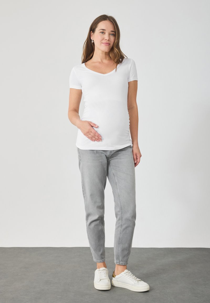 Balta, īsām piedurknēm, grūtniecību topa blūze ar V veida kakla izgriezumu, kombinēta ar gaiši pelēkiem, šauriem džinsiem un baltiem sporta apaviem. Vienkāršs dizains, gluda tekstūra.