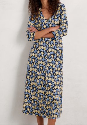 Femme aux cheveux bouclés portant une robe midi bleu marine à motifs floraux jaunes et bleu clair, debout les bras croisés.
