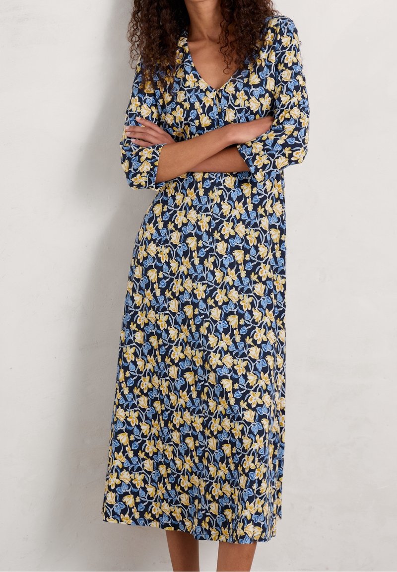 Femme aux cheveux bouclés portant une robe midi bleu marine à motifs floraux jaunes et bleu clair, debout les bras croisés.