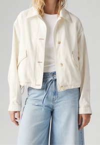 Krämfärgad cropped denimjacka med synlig sömnad, framfickor och knappstängning. Parat med ljusblå vida jeans.