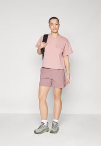 Ljusrosa kortärmad T-shirt med ficka, parat med mauvefärgade shorts. Bärs med grå vandrarskor och vita strumpor. Neutral bakgrund.