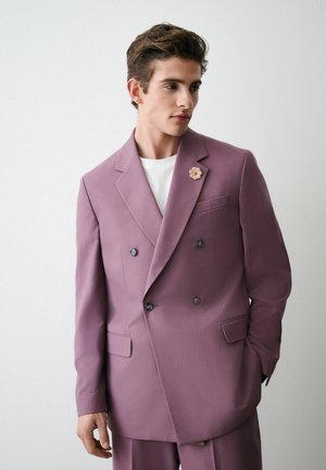 Young man wearing a double-breasted mauve suit jacket and pants, with a white shirt and a flower-shaped lapel pin, looking to the side.
