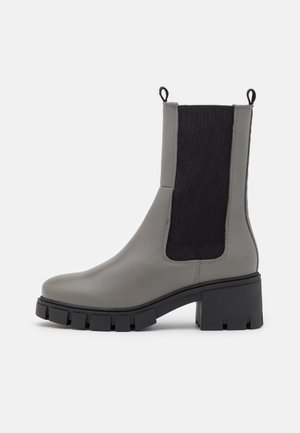 Bottine en cuir gris avec des panneaux en élasthanne noir, semelle en caoutchouc noir épaisse et languettes de tirage. Texture lisse avec un design moderne.
