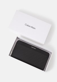 Black Calvin Klein wallet with a textured geometric pattern, zipper closure, and silver logo. Presented in a white box.