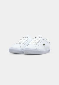 Białe skórzane trampki o gładkiej fakturze, zaokrąglonym czubku, sznurowanym fasonie oraz małym zielonym logo po boku. Przezroczysta gumowa podeszwa.