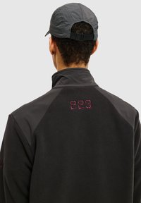 Zwarte fleecejas met hoge kraag en roze stiksels van "223" op de rug. Gecombineerd met een grijze pet, met een textuurontwerp.