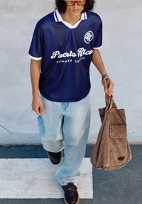 Marineblå sportsjersey med hvid krave og skrifttype, parret med lyseblå baggy jeans og mørkebrune sko. Brun tote bag i hånden.