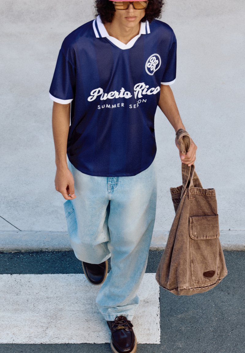 Marineblå sportsjersey med hvid krave og skrifttype, parret med lyseblå baggy jeans og mørkebrune sko. Brun tote bag i hånden.