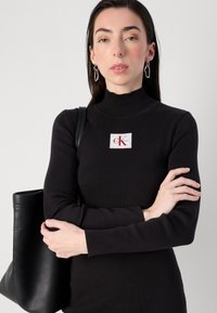 Žena s dugom kosom nosi crnu rebrastu haljinu s CK logom, drži crnu torbu i preklapa ruke, nasuprot svijetloj pozadini.