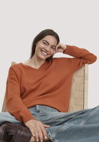 V-Ausschnitt, oversized Pullover in verbranntem Orange, aus weichem Stoff. Kombiniert mit hellblauen Jeans und dunkelbraunen Schuhen.