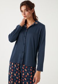 Cămașă navy cu mânecă lungă și nasturi, având un guler cu un model de turta dulce, asortată cu pantaloni care prezintă un model festiv similar.