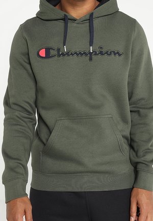 Olijfgroene Champion hoodie met voorzak en zwarte trekkoorden, gedragen door een persoon met ontspannen handen langs het lichaam.