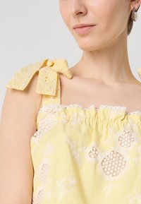 Haut sans manches jaune rayé avec des accents en dentelle et des bretelles à nouer ; présentant des motifs floraux brodés et un col froncé.