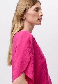 Top de color rosa brillante con un acabado texturizado, presenta mangas amplias y suavemente drapeadas, y un escote redondo, enfatizando un diseño fluido.