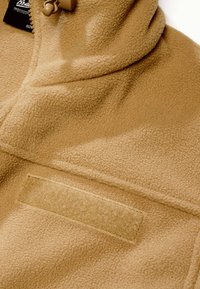 Veste polaire beige avec une fermeture éclair, un fermoir à bascule près du col et un patch Velcro horizontal sur la poitrine.