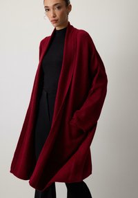 Nő laza, mélyvörös, nyitott elülső kardigánt visel fekete felső és nadrág fölött, egyszínű világos háttér előtt állva.