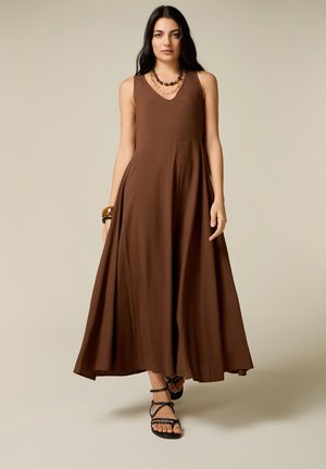 Femme aux longs cheveux foncés portant une robe maxi marron sans manches, des sandales noires à lanières, un collier et des bracelets, debout devant un fond uni.