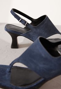 Sandales en daim bleu marine avec une bride arrière, un talon carré et un design découpé ; détaillées avec une boucle argentée et une semelle intérieure noire lisse.