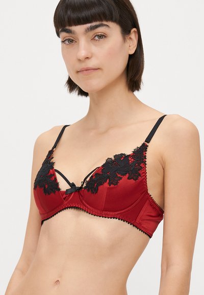 Červená saténová podprsenka s čiernym kvetinovým čipkovaným detailom, s vertikálnou výstužou, nastaviteľnými ramienkami a malou čiernou mašľou v strede.