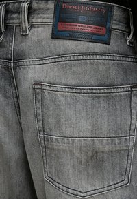 Zone de la poche arrière d'un jean Diesel gris délavé avec une étiquette rectangulaire noire, rouge et bleue indiquant la marque et les détails de qualité.