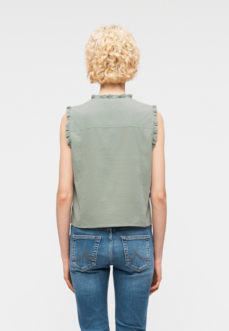 Personne aux cheveux blonds courts et bouclés, portant un haut sans manches vert sauge avec des bords à volants et un jean en denim bleu, vue de dos.