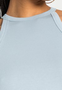 Lys blå ribbet tanktop med høj halsudskæring og diskrete syninger på skuldrene. Teksturen er glat og tætsiddende.