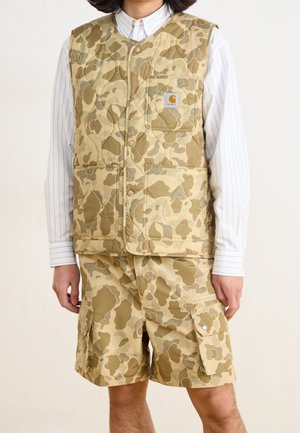 Mand iført beige og brun camouflagevest og matchende cargoshorts med hvid langærmet skjorte under mod en ensfarvet baggrund.