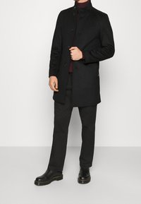Cappotto di lana nera con colletto alto, dotato di chiusura a bottone e tasche laterali, abbinato a pantaloni neri e scarpe nere.