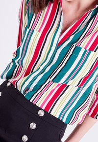 Chemise à boutons avec des rayures verticales multicolores en rouge, vert, bleu et noir ; dotée de deux poches avant et d'une texture douce et lisse.