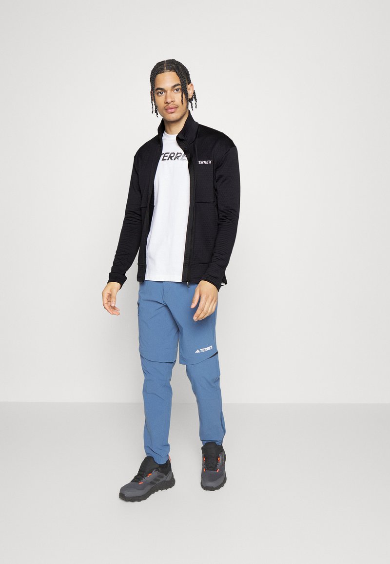 Zwarte zip-up jack met textuurpatroon, witte t-shirt, blauwe cargobroek en zwarte wandelschoenen met oranje accenten. Casual buitenkleding.